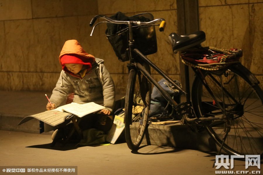 鄭州中原路京廣鐵路大橋下涵洞的馬路牙子邊，一位戴著紅色辣椒帽的小男孩趴在一個小馬扎上，上面鋪著一張車墊，在微弱的燈光下寫作業(yè)