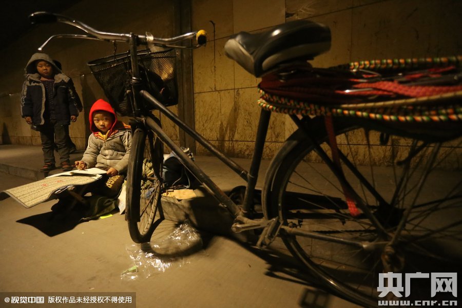 2015年11月8日晚，鄭州，7歲男孩涵洞橋下陪父母練攤，路邊燈下坐小馬扎上寫作業(yè)。